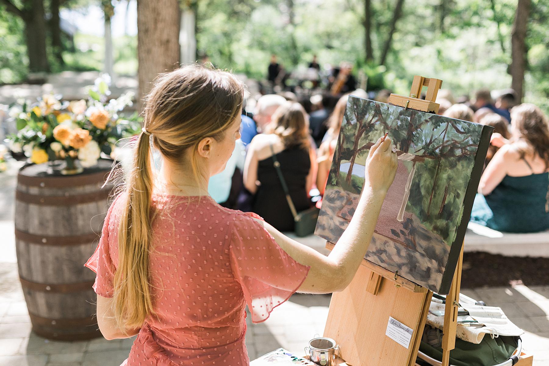 live painter artist at refined elegant outdoor wedding ceremony, with orange and yellow flowers and blue dresses, fields reserve near madison, wisconsin